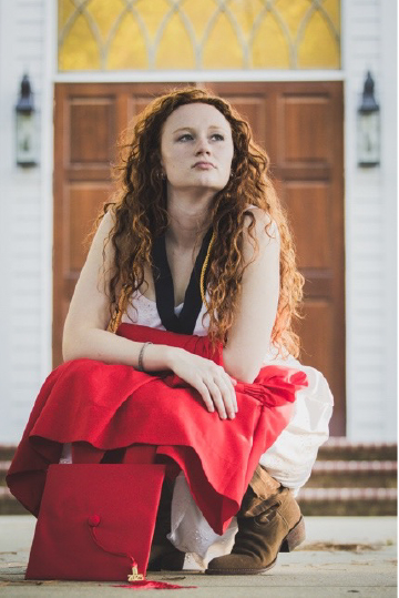 Sophia White, a first-year Honors student at Appalachian State University, sits outside holding a red graduation cap and gown while looking upward.