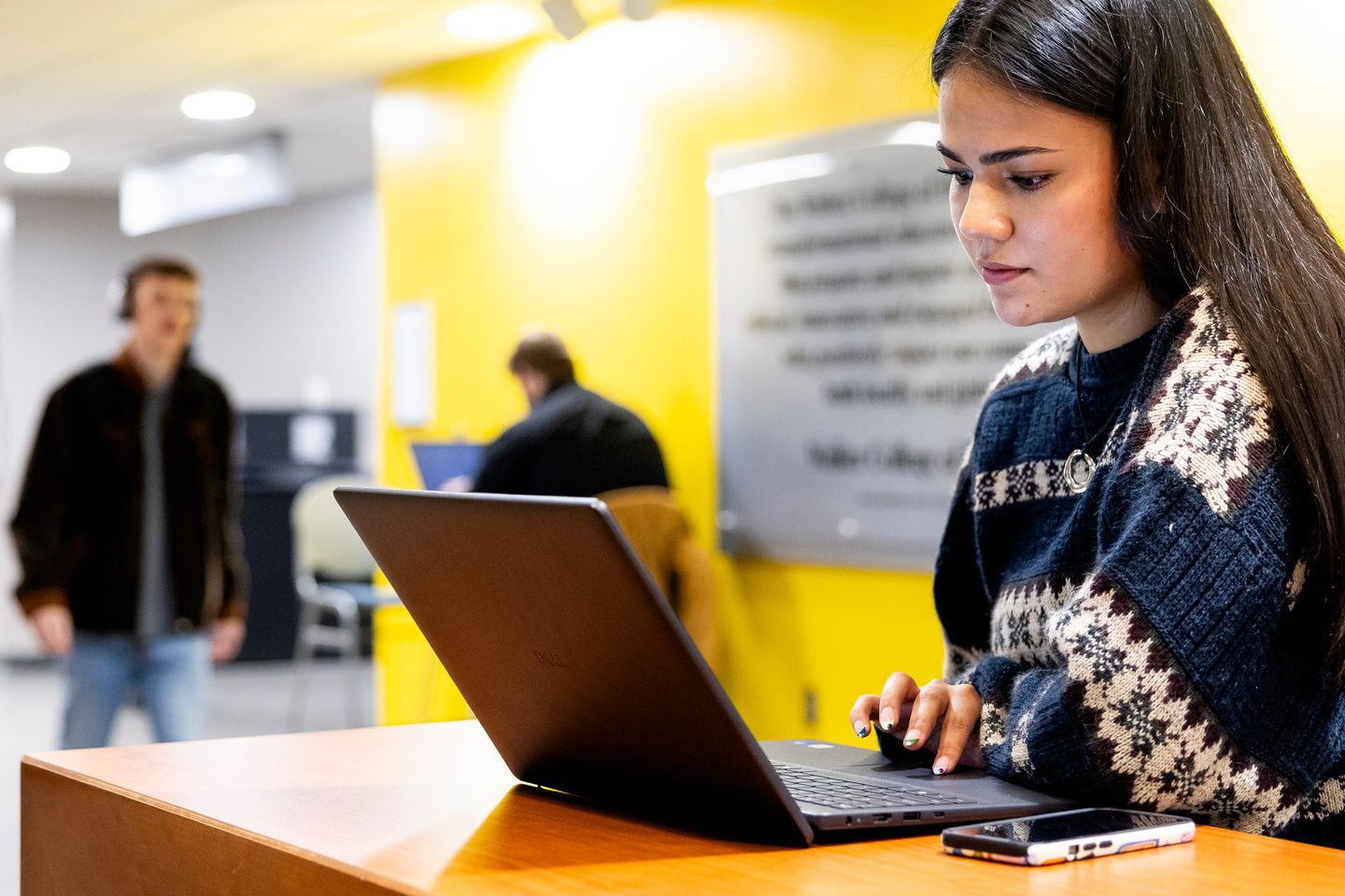 Person working on a laptop at a desk in a brightly lit indoor space with other people in the background.