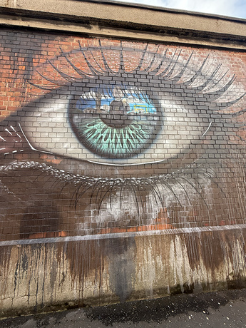 Photo of detailed painting of eye on brick wall with cloudy blue sky.