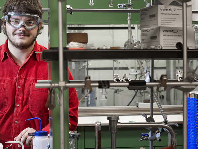 Corbin Ester working in a laboratory setting, with scientific equipment and lab supplies visible on shelves in the background.